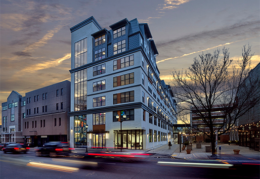 A busy city street with cars driving past an apartment building and plaza. 