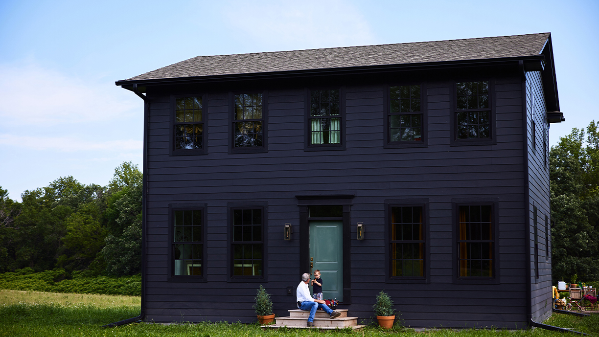 A man and young boy sit on the front steps of a black home with a green front door set in the middle of a grassy field with woods behind.
