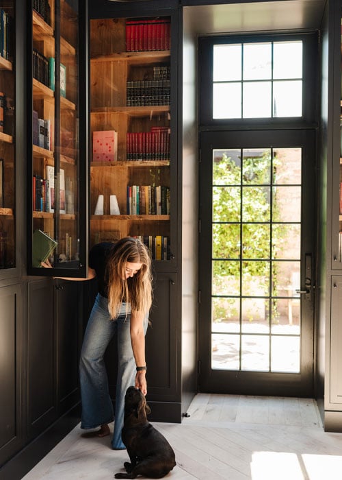 A woman unshelving a book from her bookshelf pets her dog as sunlight filters in through an adjacent door.