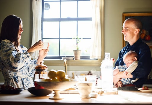 An interior shot showing light streaming through a window into a kitchen where a woman takes a photo with her phone of a man holding a sleeping baby.