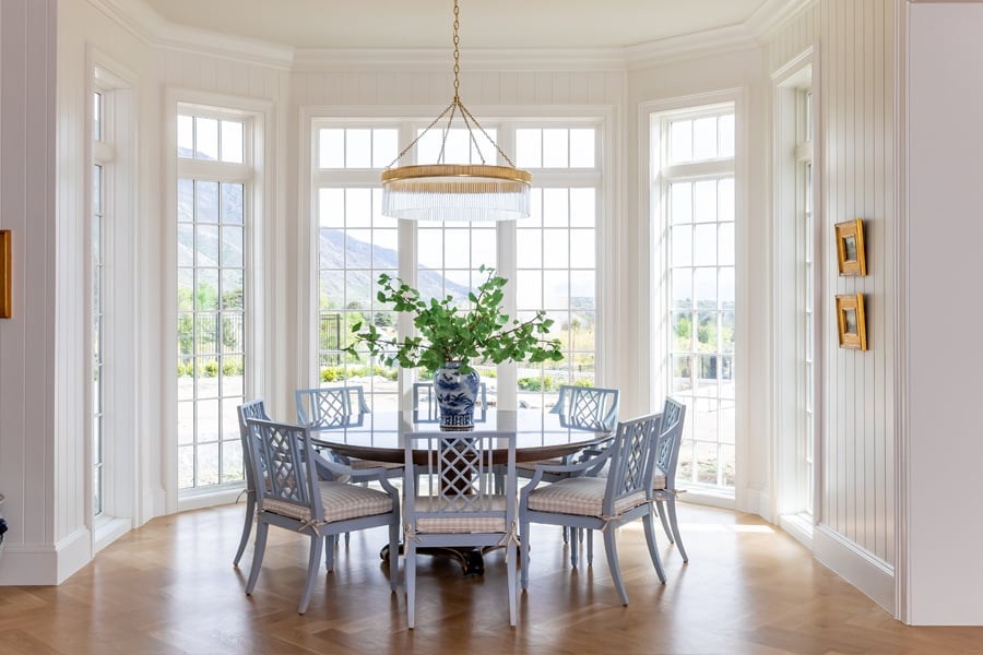 A wooden circular dining room table with blue chairs surrounded by a walk-out bay window with white grilles.