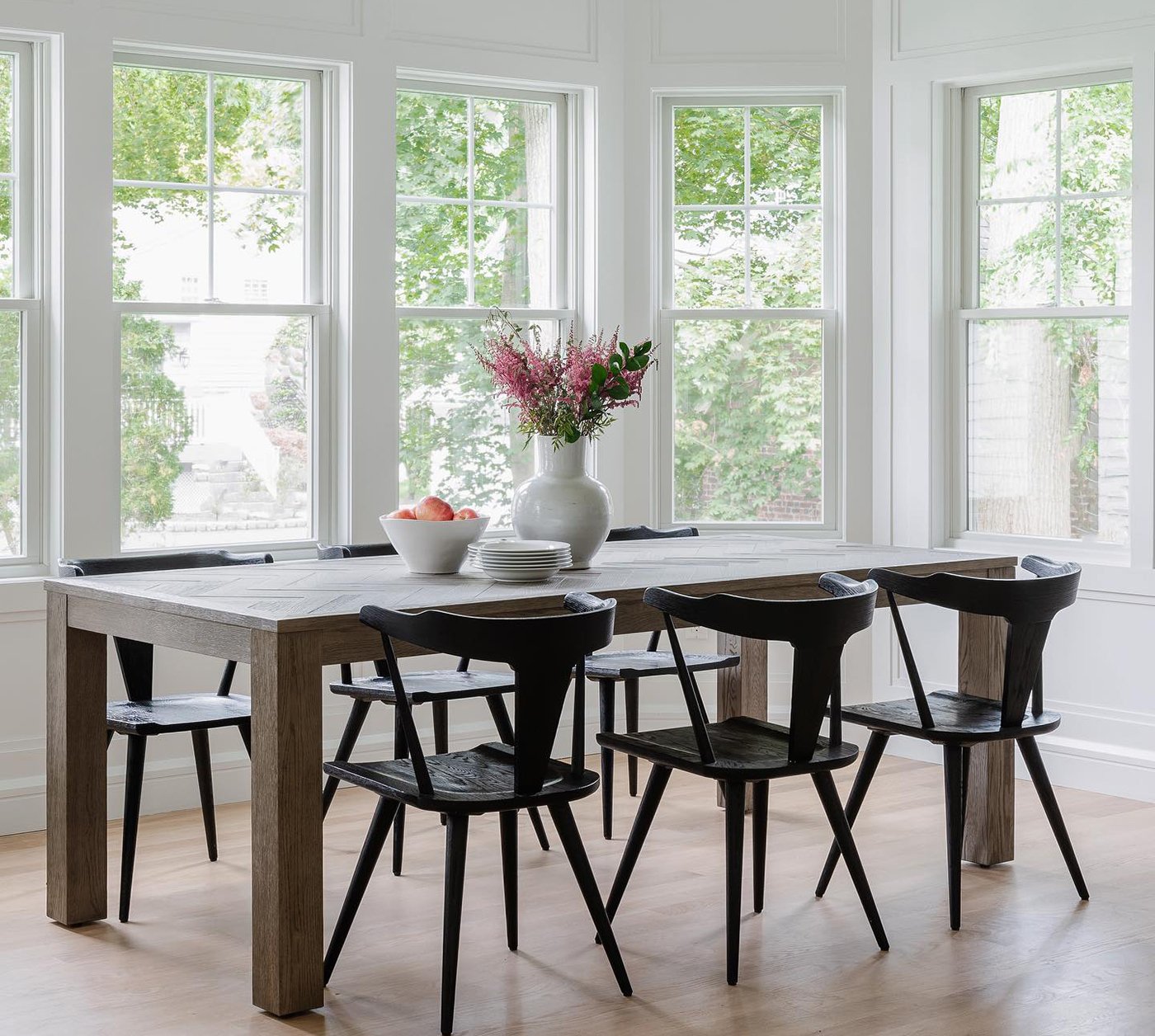 A wooden dining room table with black chairs surrounded by a walk-out bay window with white grilles. 
