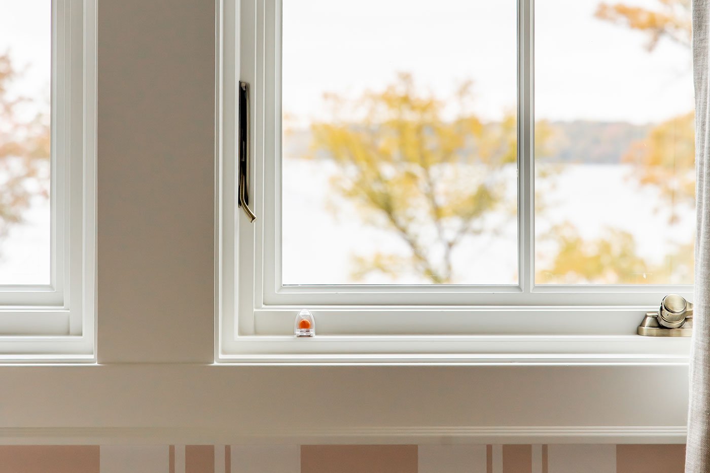 An interior shot of two white casement windows with a window opening control device and brass hardware that frame lake views. 