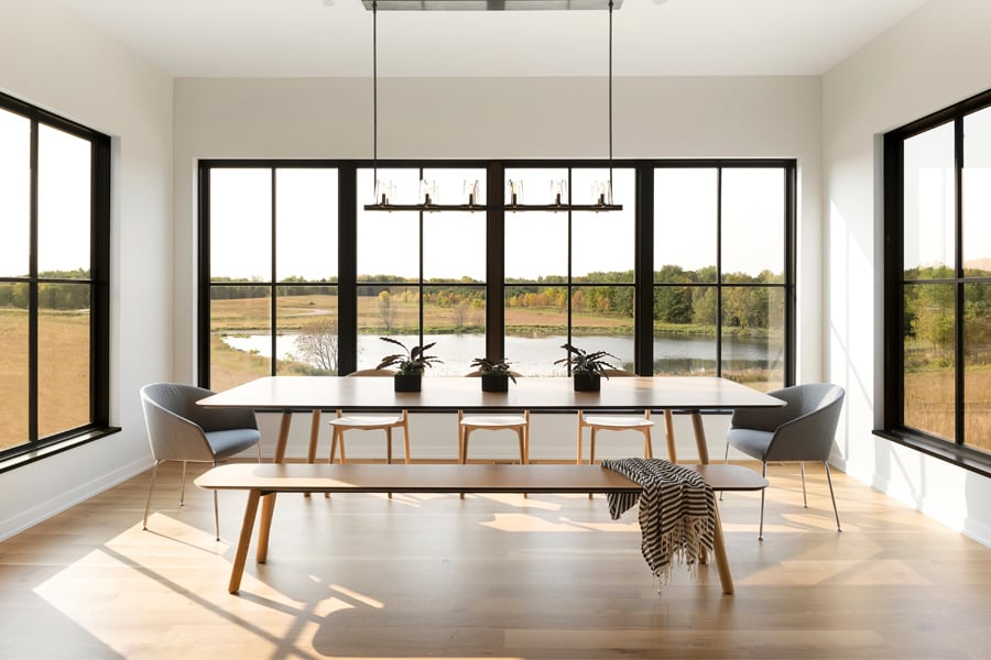 A simply furnished dining space with black windows on three sides that afford views of a prairie and wetland.