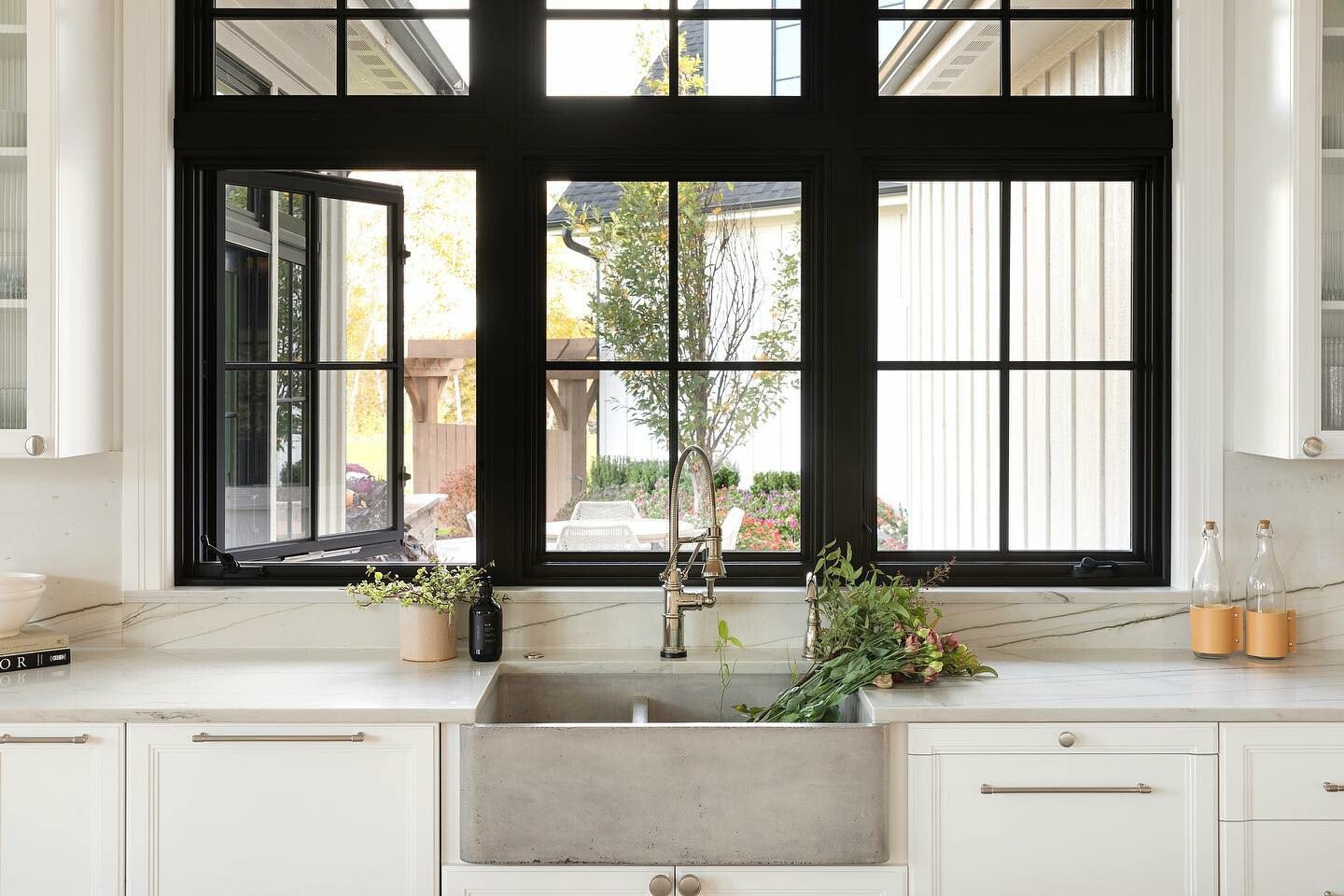 Two rows of black-framed casement windows with grilles above a concrete kitchen sink surrounded by marble countertops and white cabinetry.