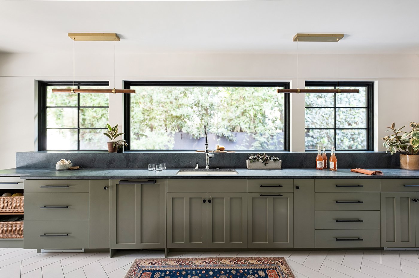 A kitchen with three picture windows above a black quartz countertop and green lower cabinetry.