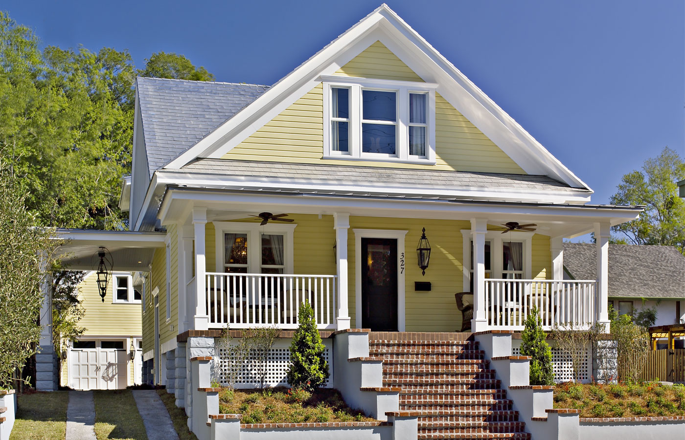 charming, two-story Craftsman-style bungalow with a bright yellow exterior and a steeply pitched gabled roof. The house features a prominent front porch supported by white columns, with a wooden railing and brick steps leading up to the entrance. The porch is adorned with comfortable seating, and there are two large windows flanking the front door. The landscaping is minimal, with a well-kept lawn, small shrubs, and a simple walkway leading to the house. The home is set against a backdrop of trees, adding to its welcoming and cozy atmosphere.