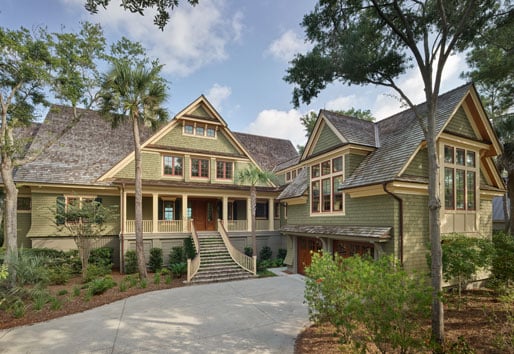 large, two-story Craftsman-style bungalow surrounded by lush greenery and tall trees. The house features a steeply pitched roof with shingles, multiple gables, and an inviting front porch with a wide staircase leading up to the entrance. The exterior is a mix of wood and stone, with numerous windows allowing natural light to pour into the home.  