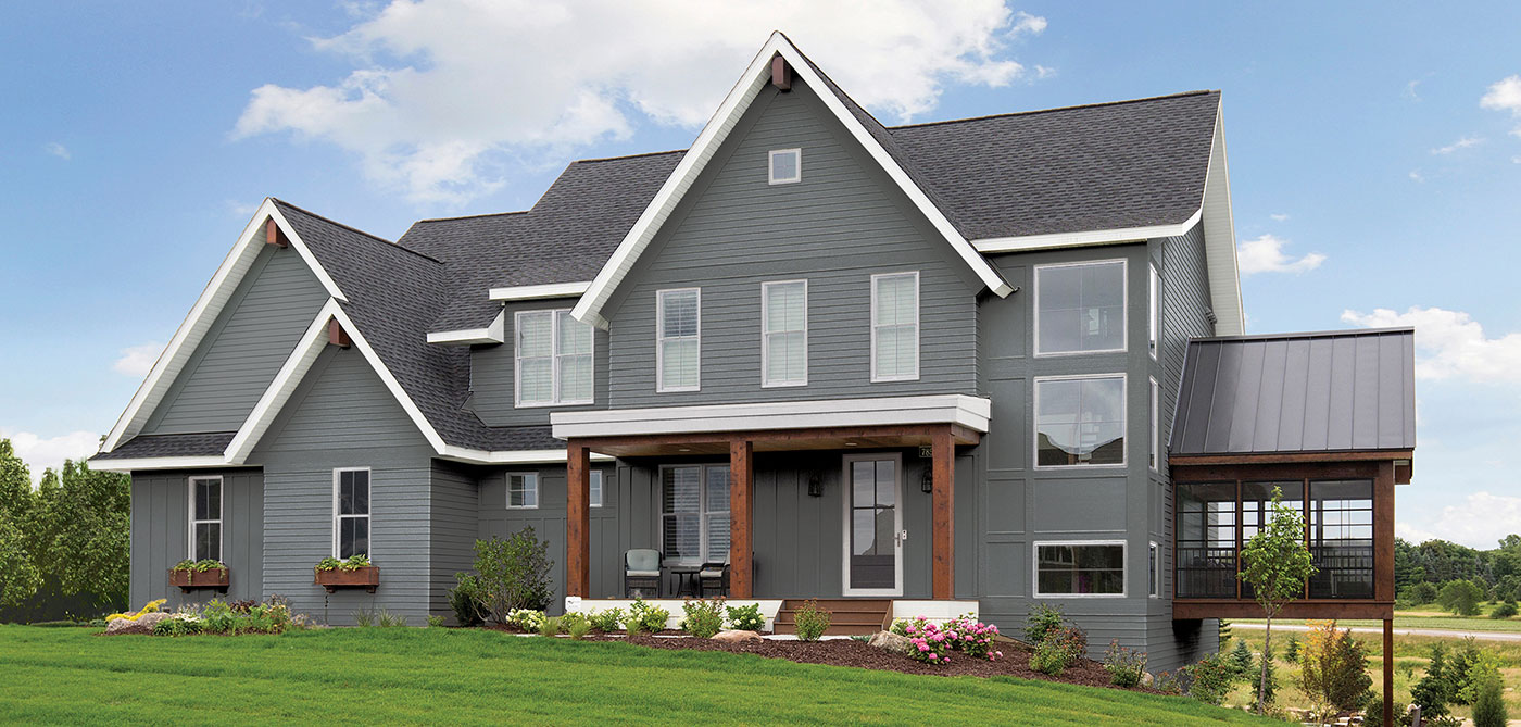 large blue-grey, two-story, modern farmhouse positioned centrally on a large green lawn. The porch has wooden columns and steps leading up to a single front door. There’s a wood trimmed sunroom located on the right of the image. This house hosts a beautiful combination of double pane and fixed windows in white.