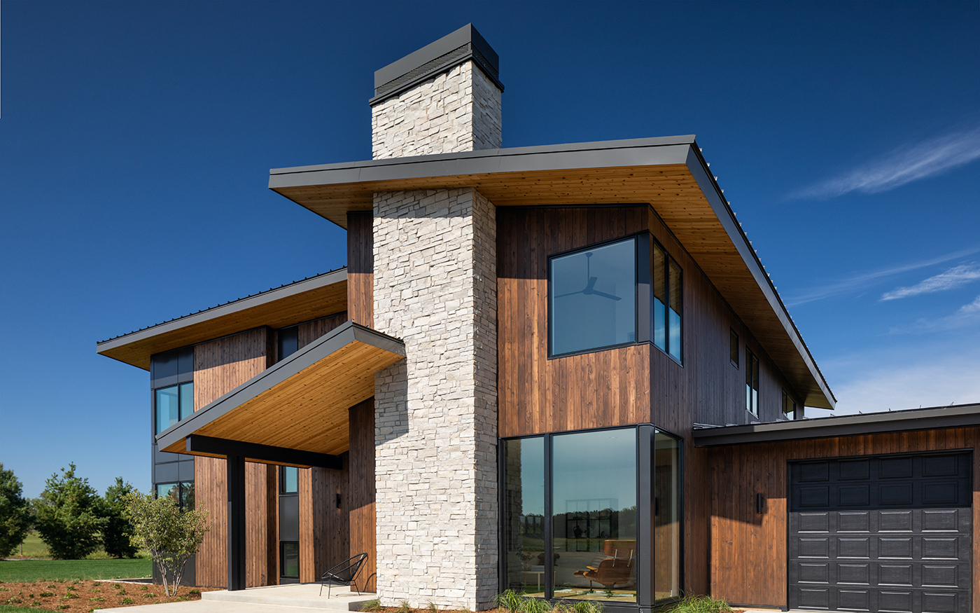 A modern house with wood paneling, gray stone chimney, black garage door and E-Series Windows with black frames. 
