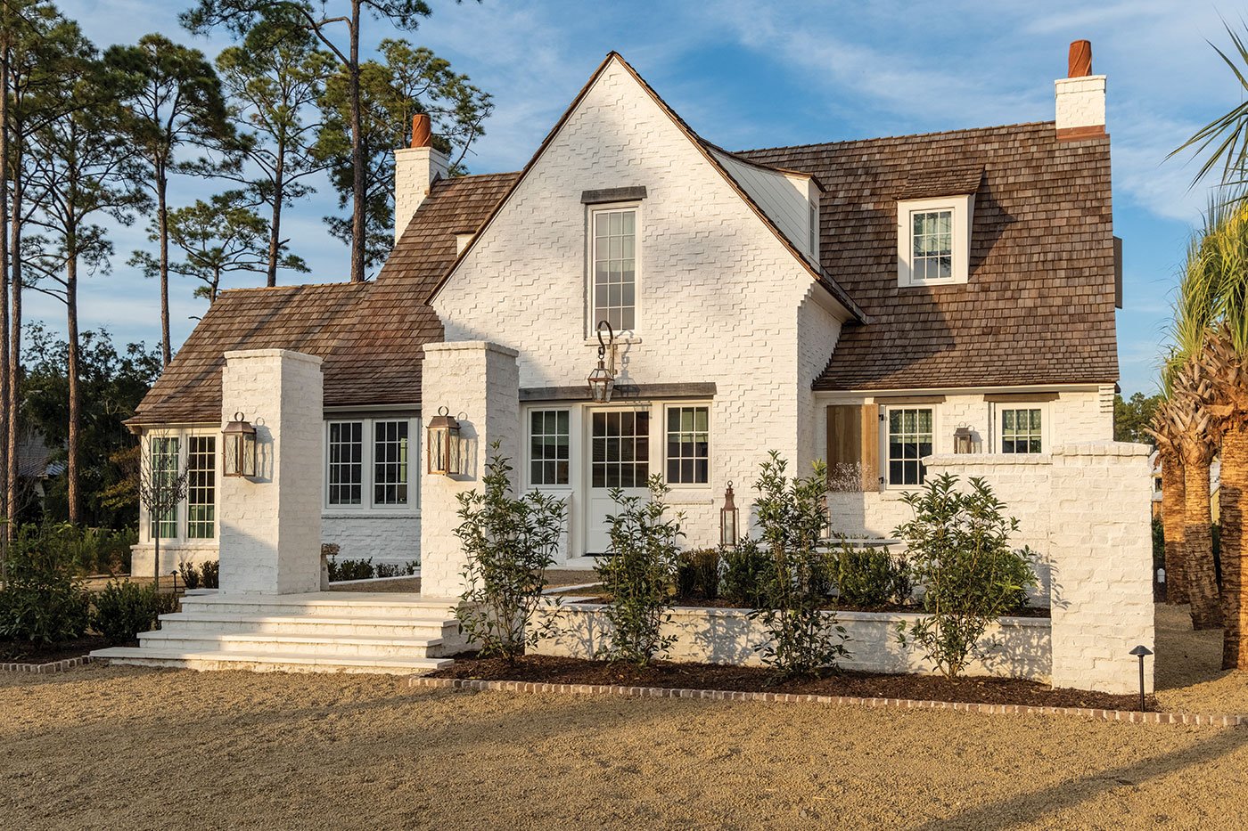 An exterior view of cottage with a white painted brick exterior and a cedar shake roof complete with dormers.