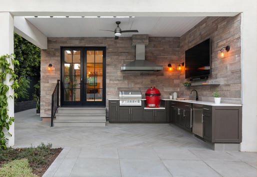 An outdoor kitchen with a grill, smoker, range hood, wall mounted TV, countertops, sink, and cabinetry.