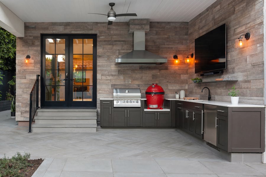 An outdoor kitchen with a grill, smoker, range hood, wall mounted TV, countertops, sink, and cabinetry.