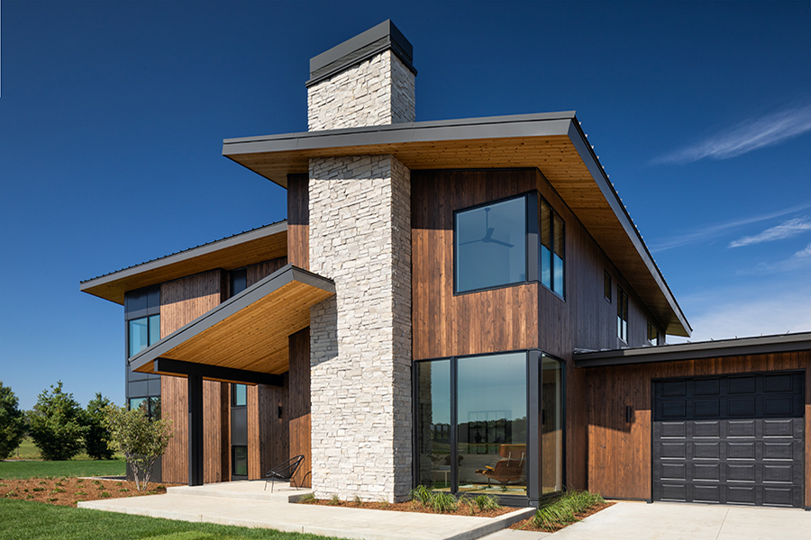 A modern home with wood paneling, a gray stone chimney, and black picture windows.