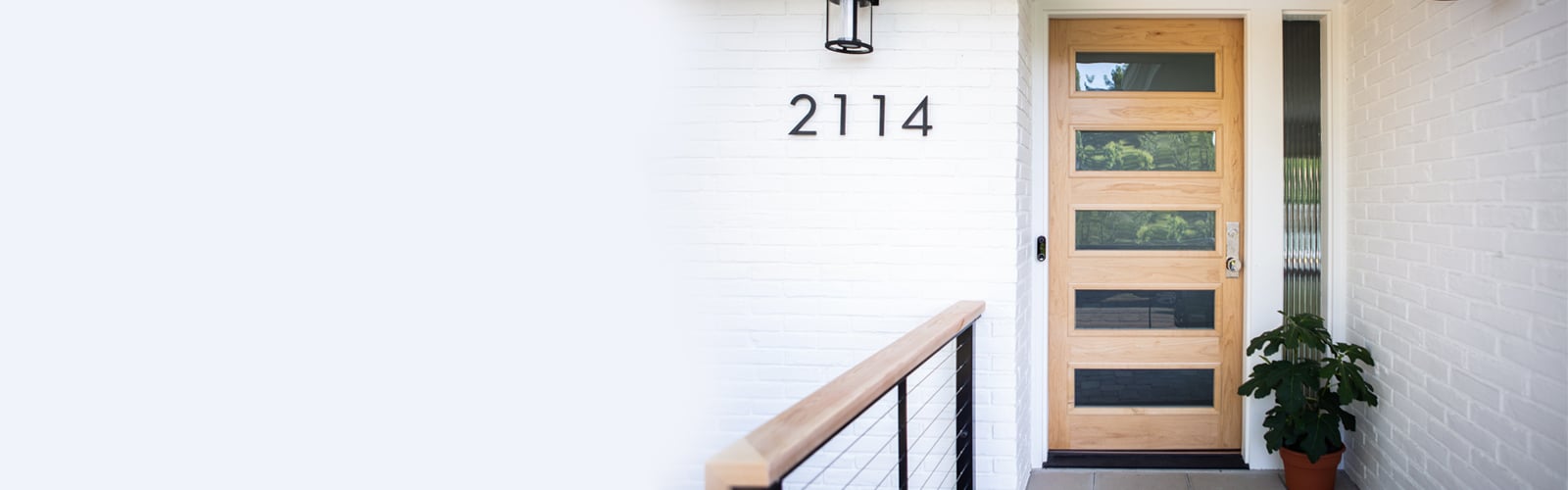 A modern front door with a light-colored wood exterior and horizontal glass panels popping against the white brick walls of a Mid-Century Modern home.