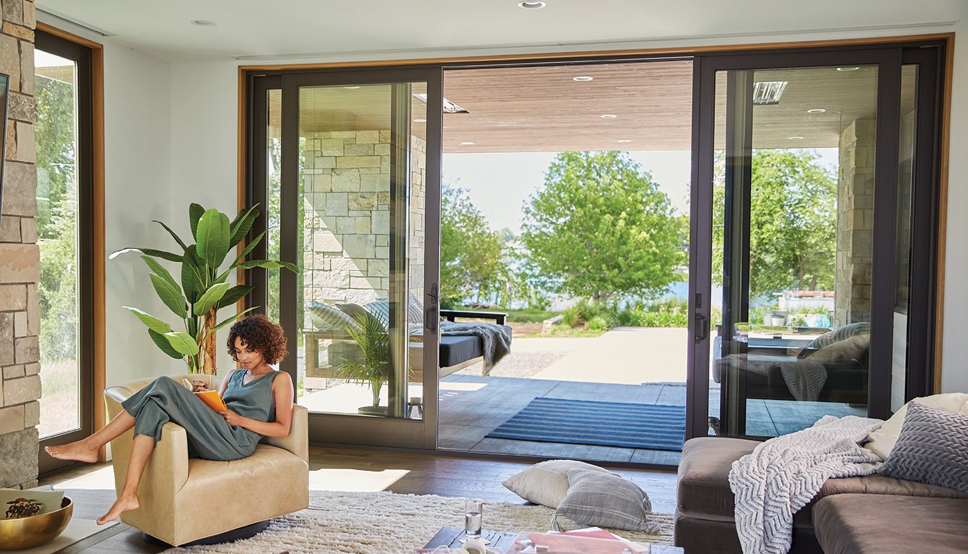 A woman reads in a chair while behind her a bi-parting A-Series Gliding Patio Door stands open to a lake view.