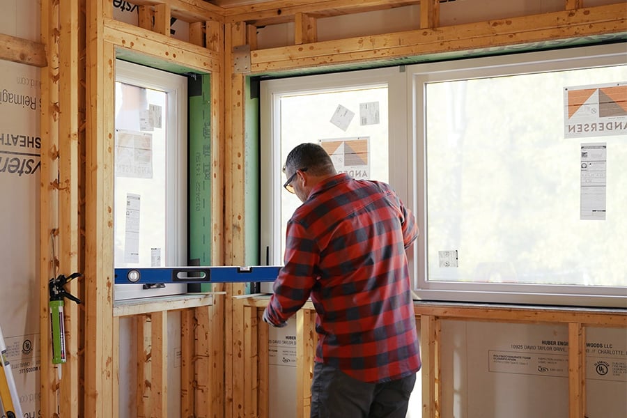 A man in a framed-out new construction home uses a level to check that a window’s properly installed.