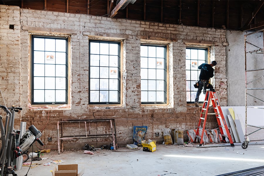 Inside a building with exposed brick walls, a man installs the last window in a row of four black-framed casement windows with grilles.  