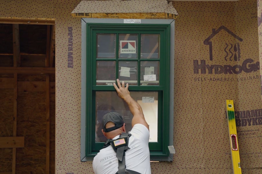 A man inserts a green-framed casement window with modified colonial grilles into a rough opening.