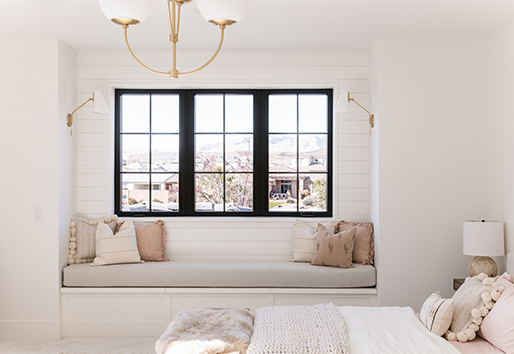 A bedroom with a neutral color scheme, shiplap walls, and three black-framed windows with a window seat underneath.