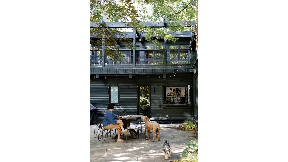 An exterior photo of a home showing a woman sitting at a table on a patio, two dogs milling about, and inside an open window, a man mixing a cocktail. 
