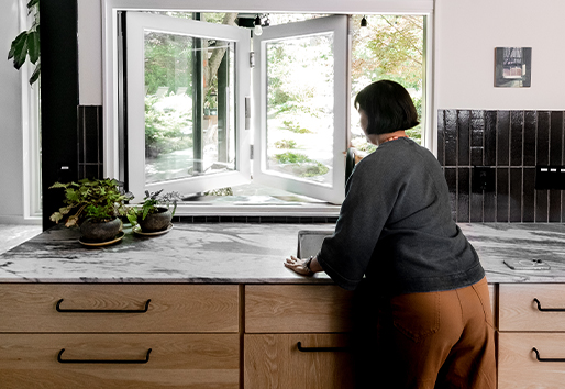 A woman reaches over a marble countertop and kitchen sink to open a folding pass-through window.