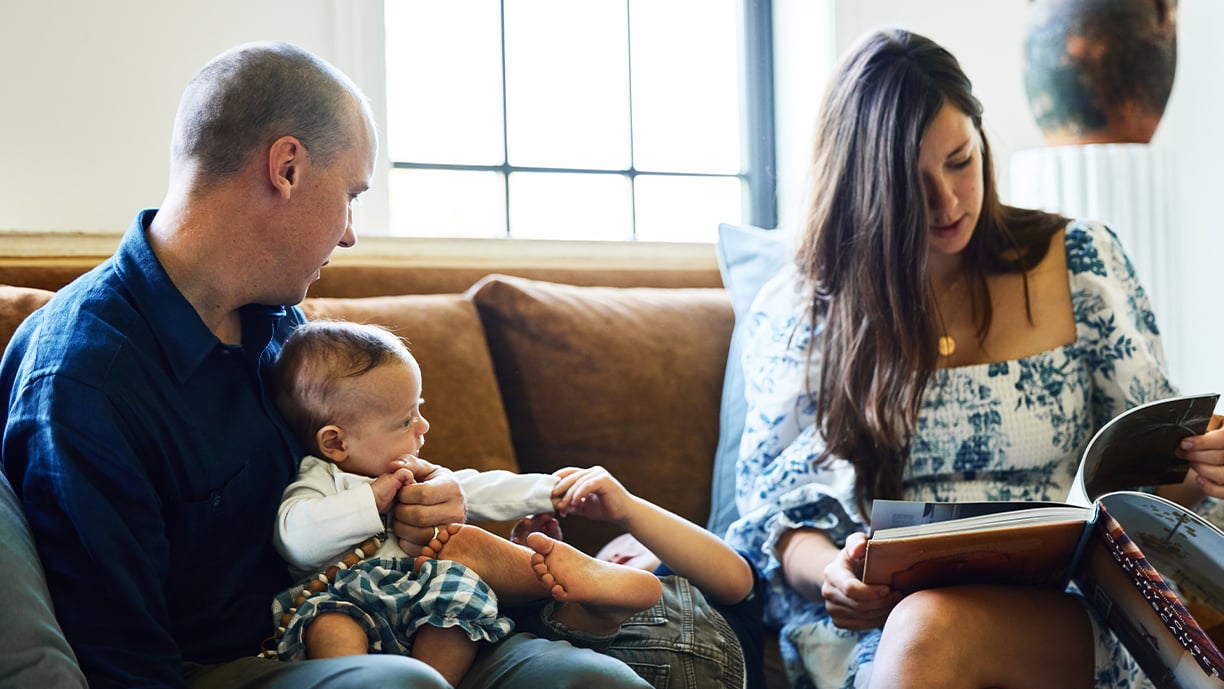 Light streams in the window of a living room where a man holding a baby sits on a brown velvet couch next to a young child and a woman reading a book. 