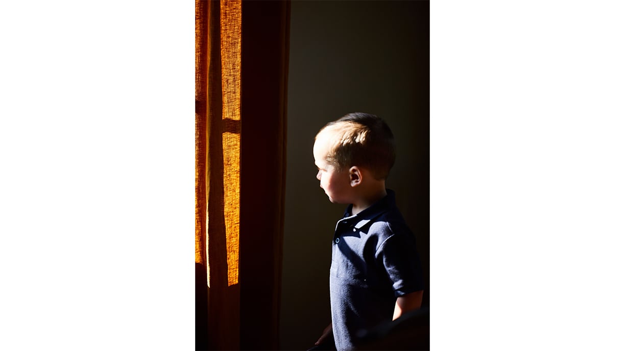 A young child wearing a navy-blue collared shirt looks out a window framed by ochre linen curtains.