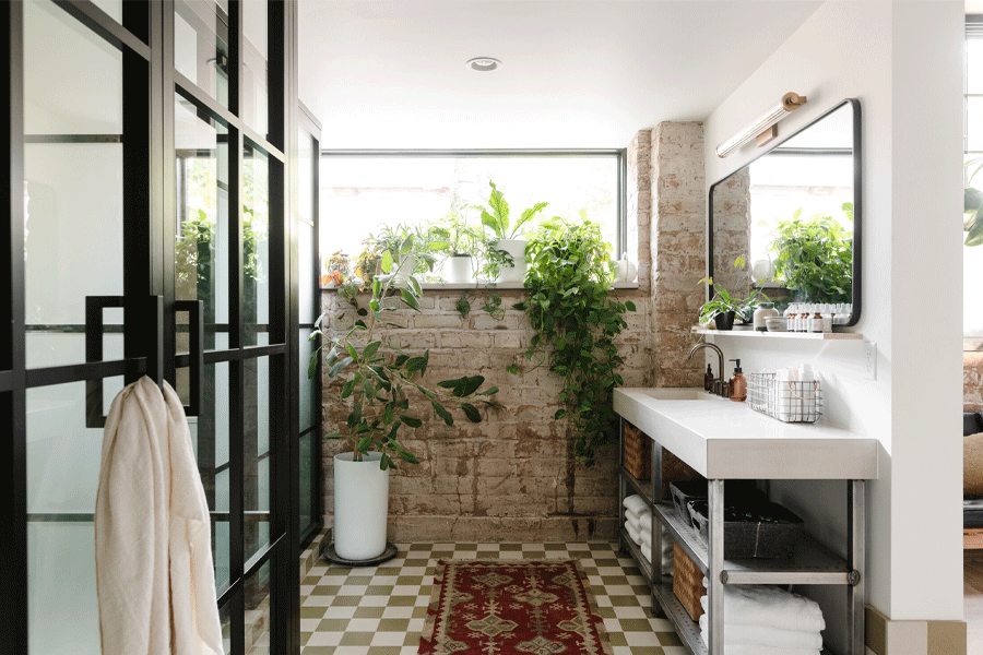 A bathroom with a green-and-white checked tile floor, black steel shower enclosure, and big window full of potted plants. 
