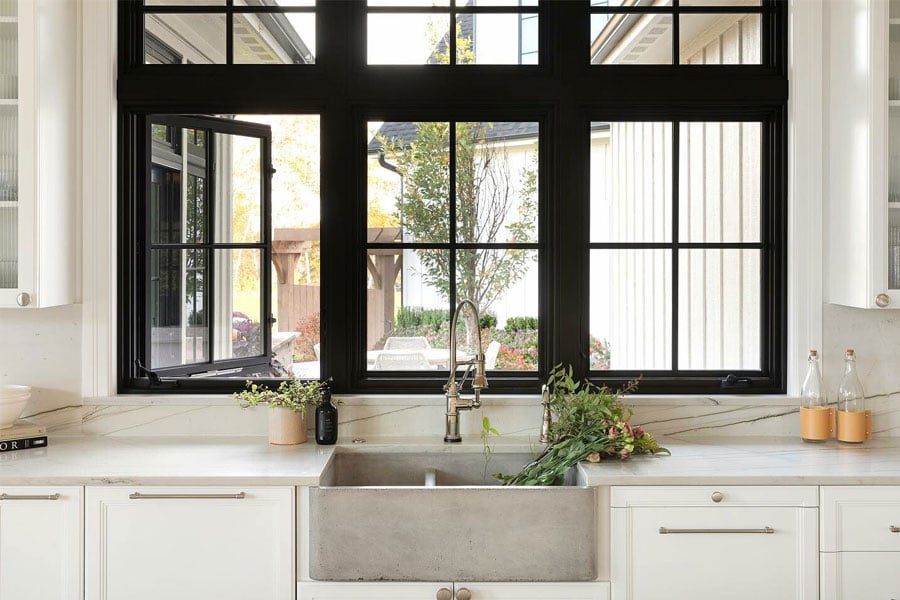 Two rows of black-framed casement windows with grilles above a concrete kitchen sink surrounded by marble countertops and white cabinetry.