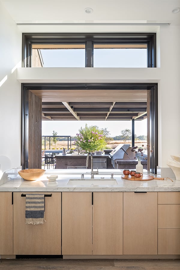 An open pass-through window connects an indoor kitchen with white oak cabinetry and marble countertops with an outdoor kitchen shaded by a large overhang.