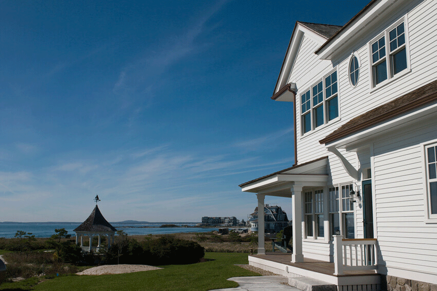 A white colonial home with coastal windows faces a gazebo surrounded by sea grass and the ocean beyond