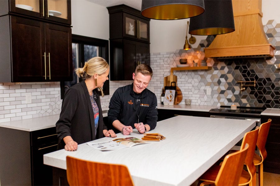 A homeowner and contractor stand at a kitchen island discussing options for a patio door replacement with black cabinetry and black-and-white tile backsplash in the background.