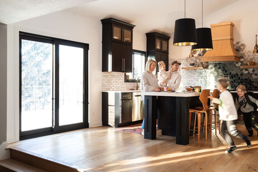 Parents laugh while their children race around a kitchen featuring black cabinetry and a black framed sliding glass door. 