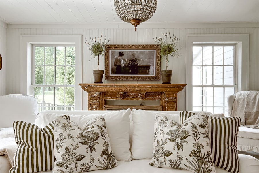 A white couch with two white windows behind and a weathered wooden fireplace mantle.