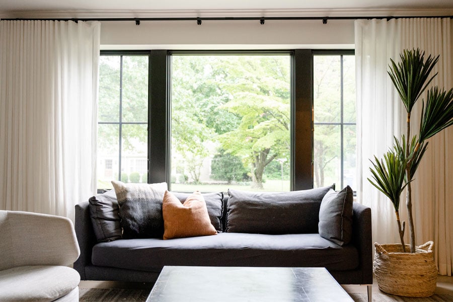 A living room couch sits in front of a picture window flanked by two casement windows with grilles and hung with white curtains. 