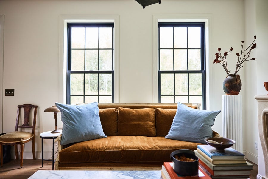 A white living room with a brown velvet couch in front of two black-framed single-hung windows.