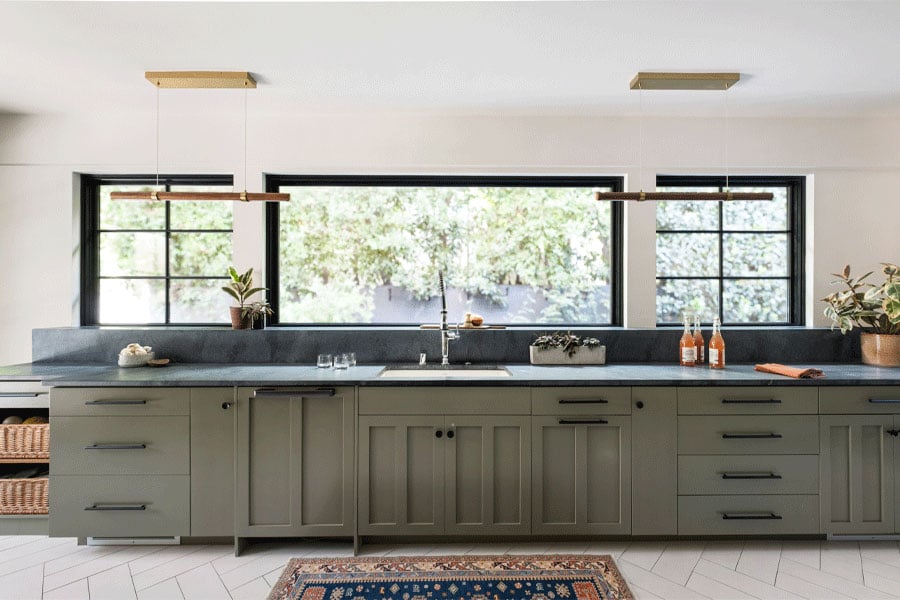 A kitchen with green cabinetry and black countertops has a view to the outdoors through three black-framed Andersen picture windows.
