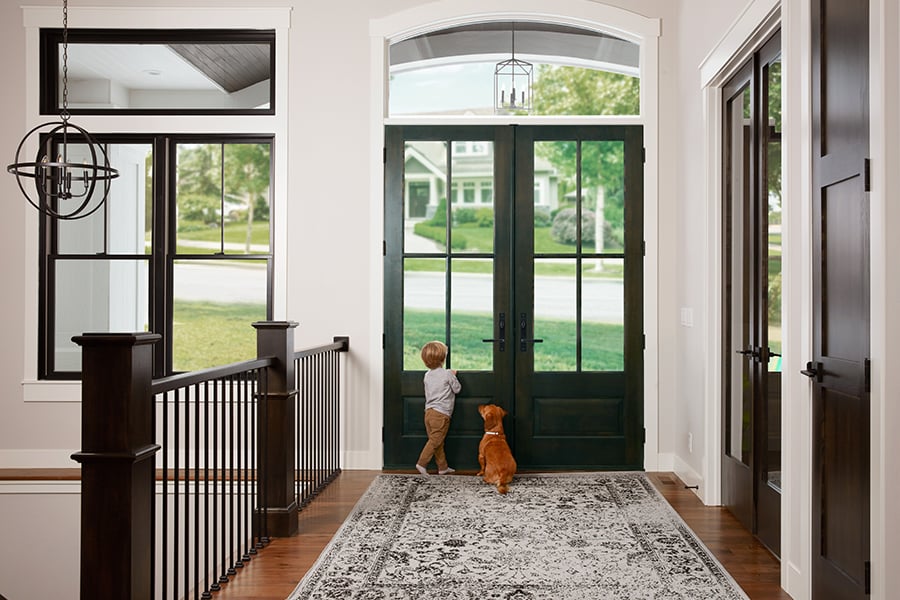 A small child and dog stand in a front hall with wood floors and a rug and look outside through a set of double front doors.