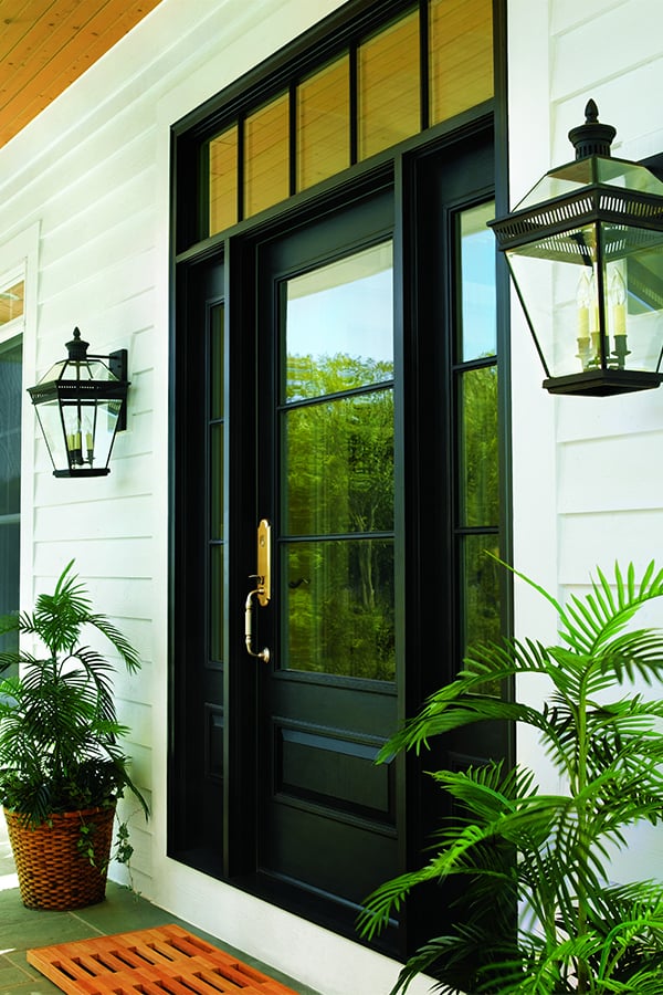 A entryway with a black front door featuring a large glass panel with a transom window above, sidelights on both sides, and potted plants and lantern-style lights flanking the door, which is on a white house.
