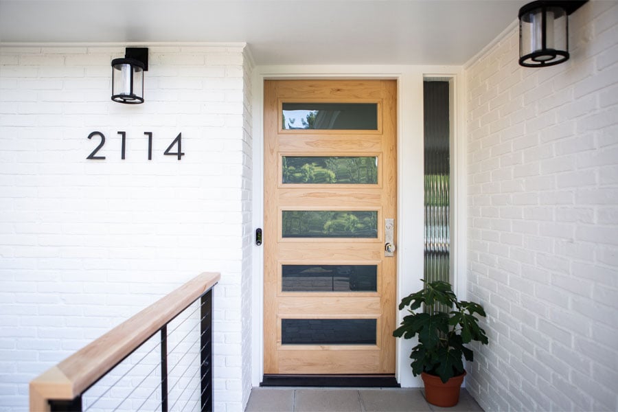 A natural wood front door with five horizontal windows in it and a potted plant next to it makes a beautiful entrance to a white-painted brick home. 