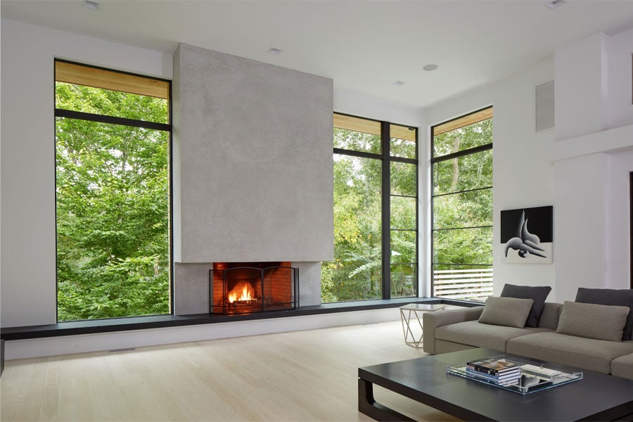 A modern living room with monumental concrete fireplace flanked by black-framed floor-to-ceiling windows and a white oak wood floor.