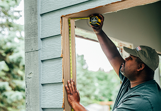 A man measures a rough opening inside a home with green wood siding. 