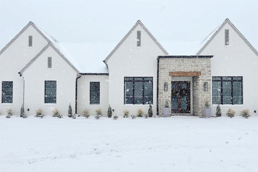 A white home with black windows and stone entryway is cozy against the snowstorm outside.