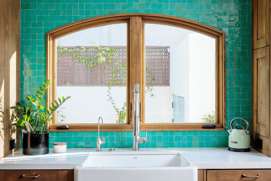 Two arch-top casement windows above a kitchen sink with a turquoise zellige tile backsplash and alderwood cabinetry.