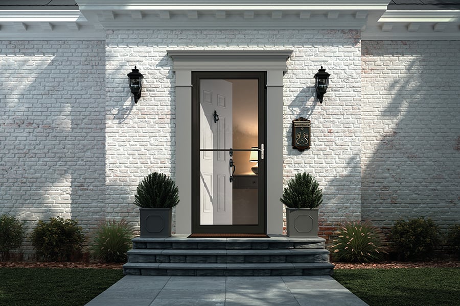 An exterior view of a home’s entrance with white brick siding, a dark bronze storm door, and gray trim framing a white front door.