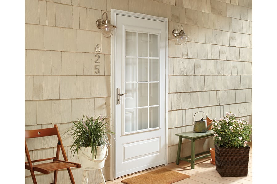 An exterior view of a home’s entrance with cedar shake siding and a white front door with a white ¾ Light Panel Ventilating Storm Door surrounded by potted plants, a bench, and a chair.
