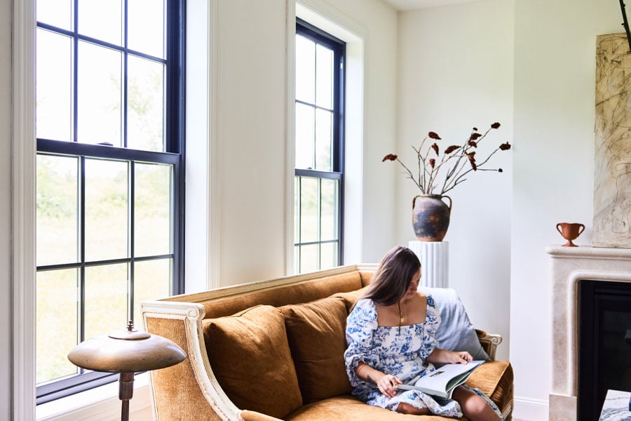 Two black windows with colonial grilles bring a flood of natural light into a living room where a woman reads a book on the couch.