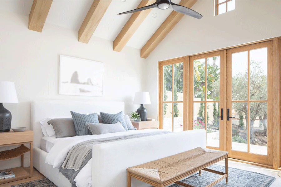 A bedroom with white walls and white oak Andersen French doors, ceiling beams, and furniture has a view of the garden outside.