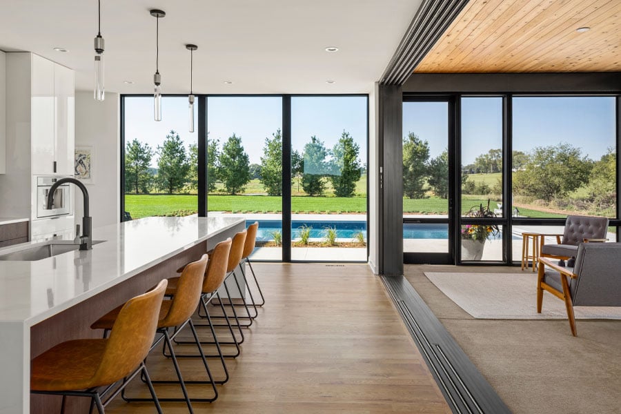 A MultiGlide™ door connects a kitchen and porch creating a large living space with floor-to-ceiling windows facing a swimming pool.  